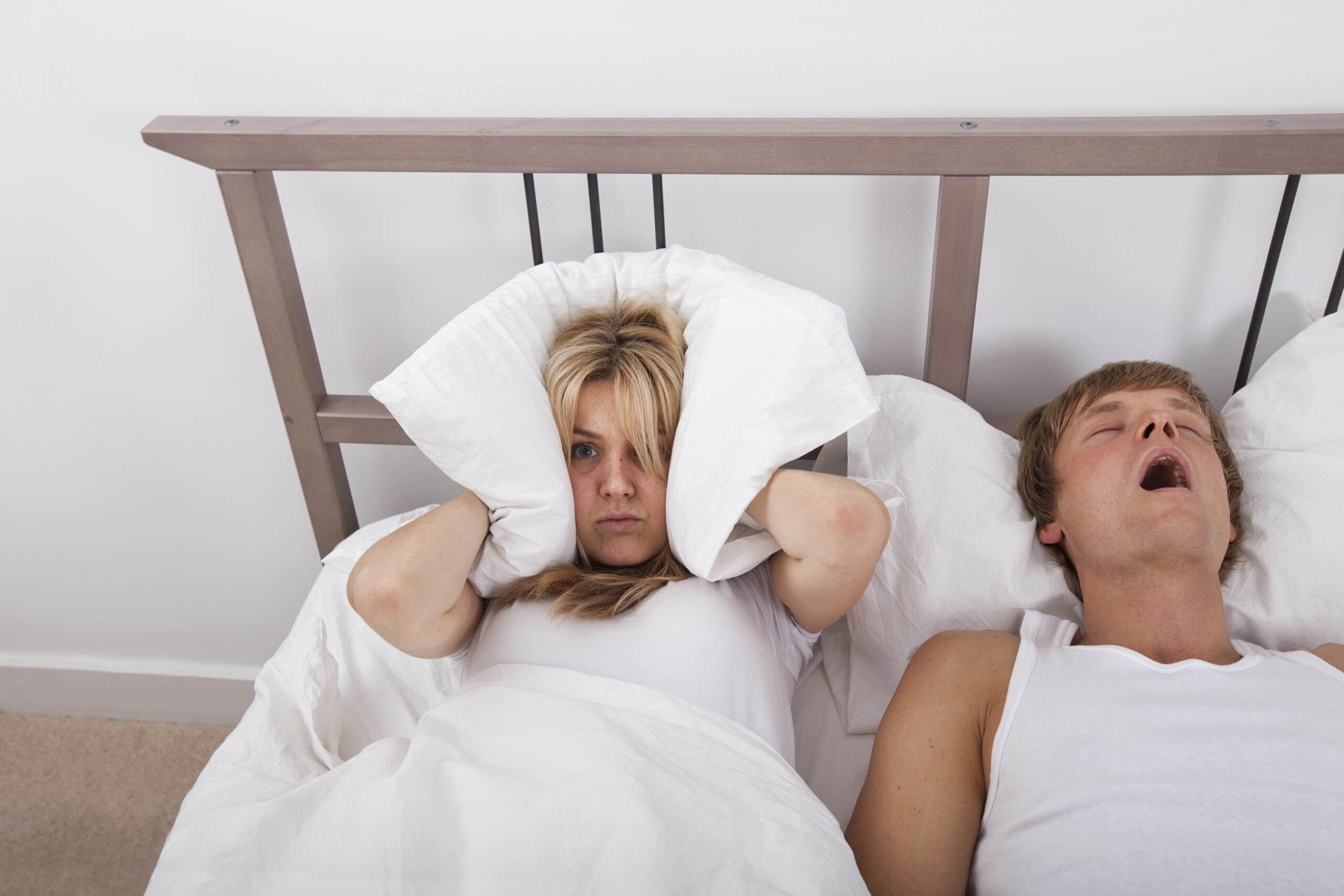 Frustrated woman covering her ears with a pillow whilst partner is beside her asleep and snoring