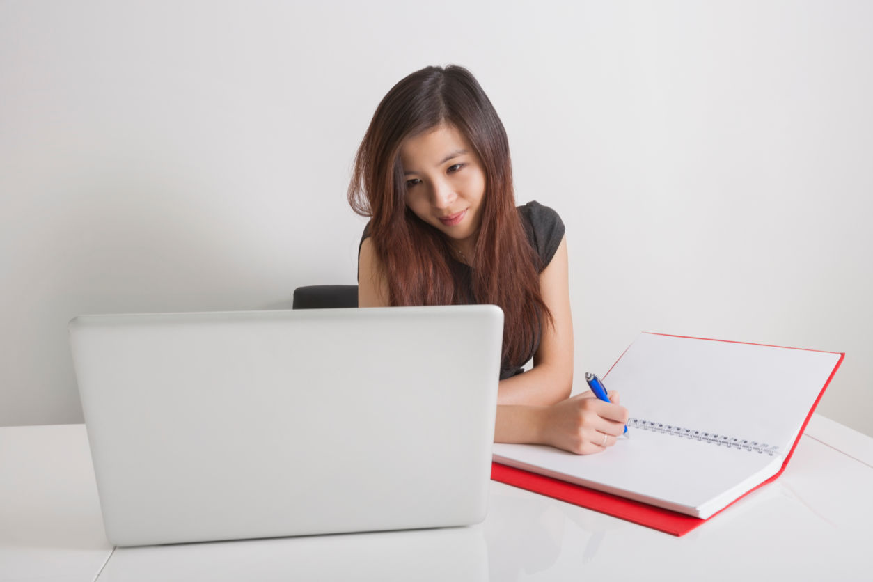 Businesswoman writing in red diary while looking at a white laptop in office whilst working freelance jobs
